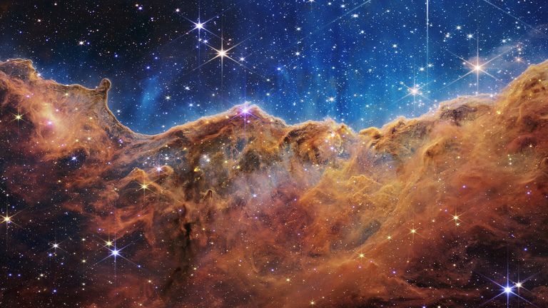 The "cosmic slopes" From the Carina Nebula in an image horizontally divided by a wavy line between a nebula-forming cloudscape along the lower portion and a relatively clear upper, with data from NASA's James Webb Space Telescope 